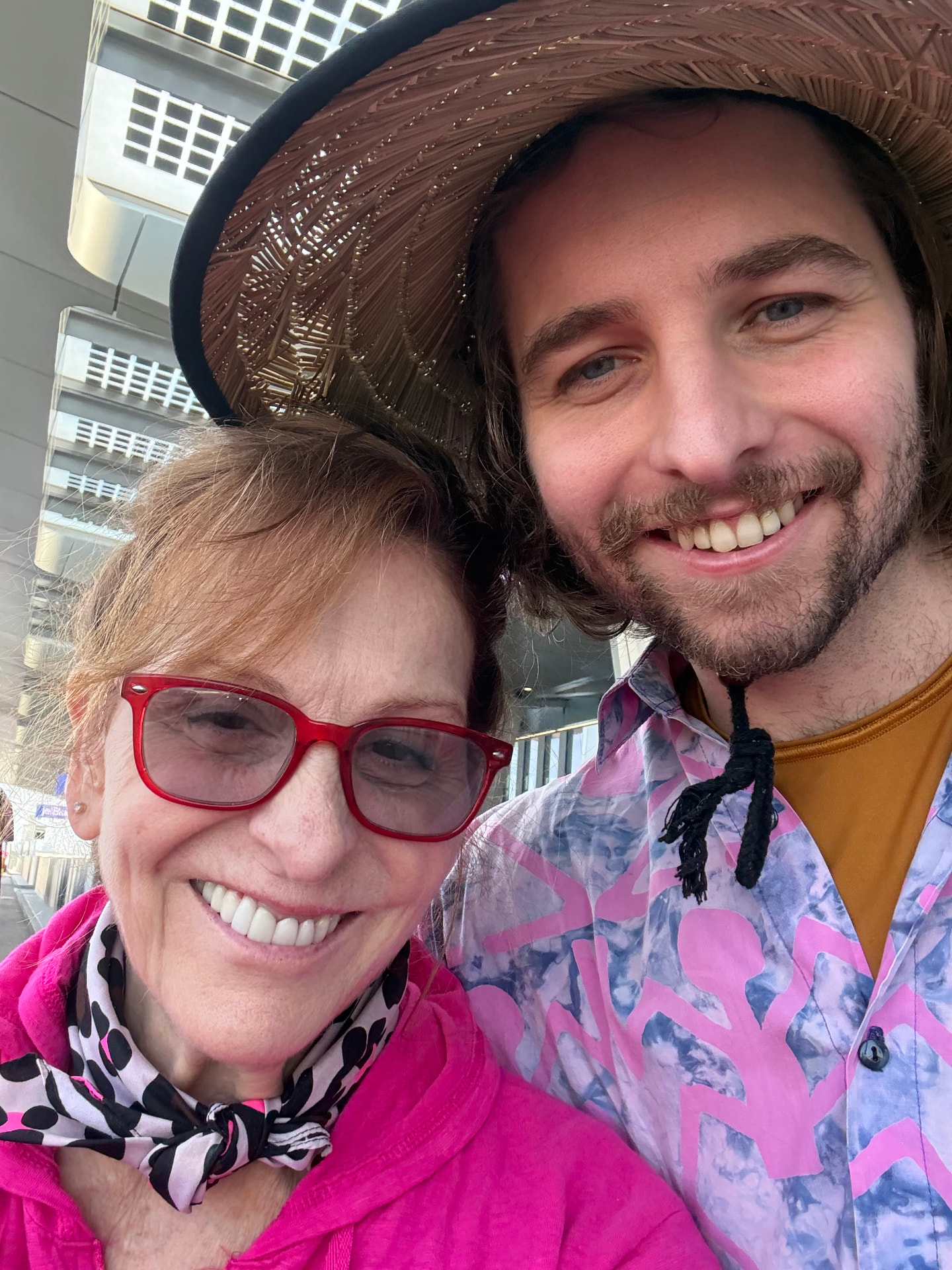 Michele and Dylan Finley smiling together outdoors, Michele in bright pink with red glasses and Dylan in a straw hat